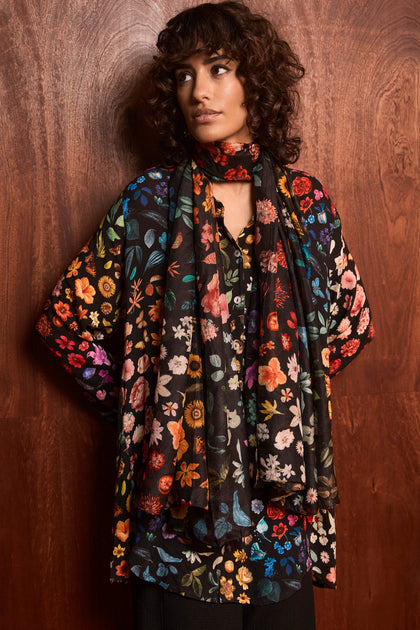 A woman with curly, shoulder-length hair and a neutral expression stands against a wooden wall, wearing a black botanical print top and the Midnight Garden Silk Scarf.