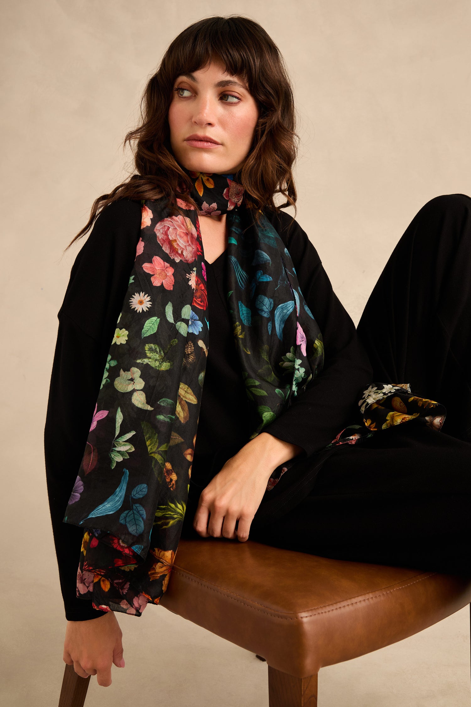 A woman sits on a brown chair against a beige background, wearing a black outfit and the Midnight Garden Silk Scarf—a black silk scarf featuring a vibrant botanical floral and bird pattern—while looking to the side.