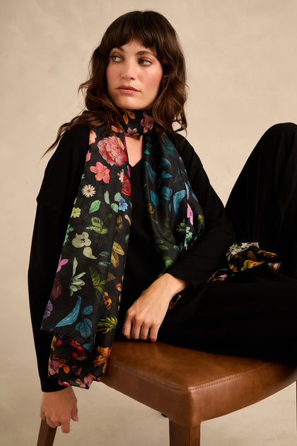 A woman sits on a brown chair against a beige background, wearing a black outfit and the Midnight Garden Silk Scarf—a black silk scarf featuring a vibrant botanical floral and bird pattern—while looking to the side.