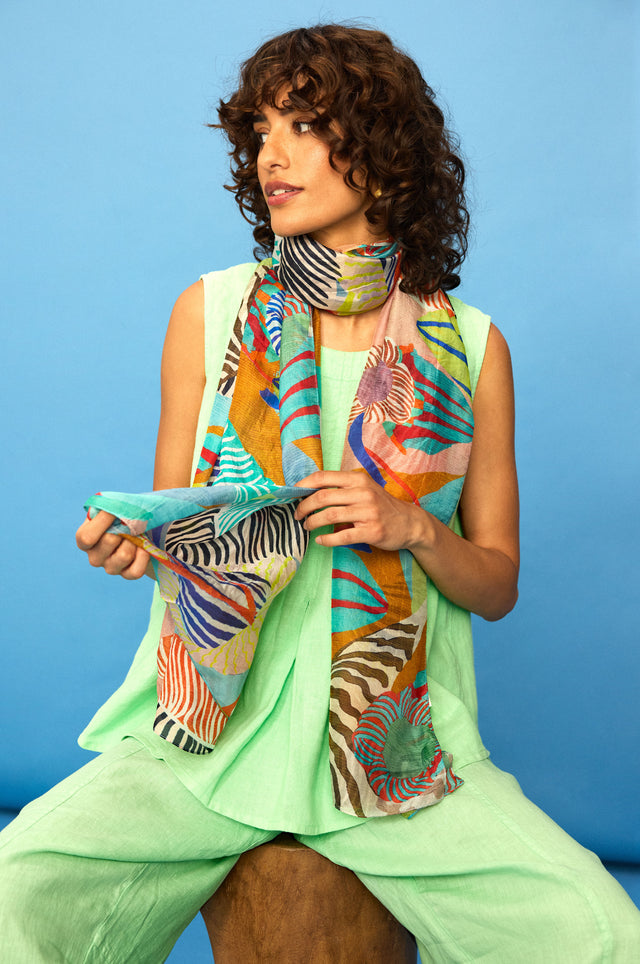 A woman with curly hair wears a light green outfit and the Tropical Orchid Fantasy Silk Scarf while seated on a wooden stool against a blue backdrop.
