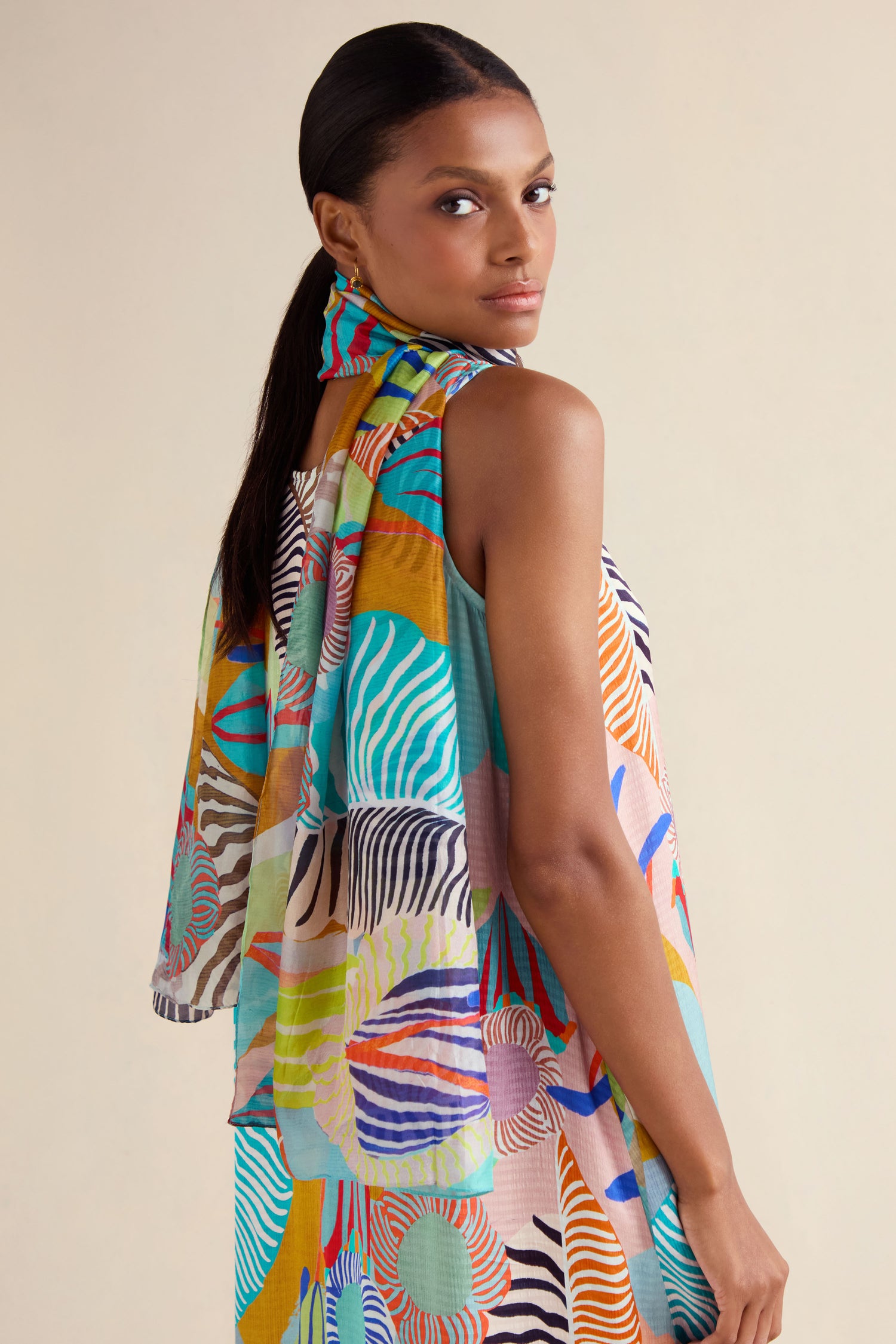 A woman with long dark hair wears a colorful dress with abstract patterns and the Tropical Orchid Fantasy Silk Scarf, looking over her shoulder at the camera against a plain background.