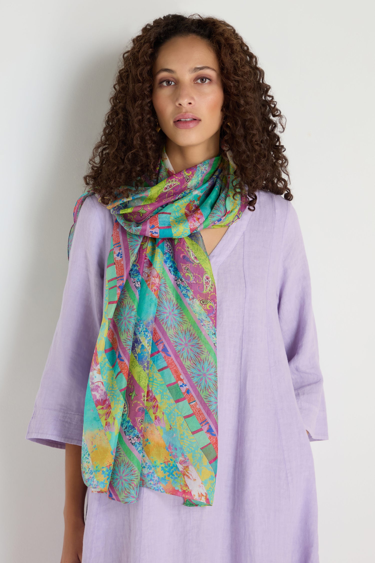 Woman with curly hair wearing a light purple dress and the Archival Stripe Silk Scarf, featuring a colorful stripe design, standing against a plain white background.