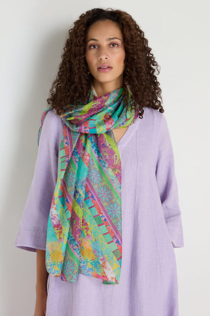 Woman with curly hair wearing a light purple dress and the Archival Stripe Silk Scarf, featuring a colorful stripe design, standing against a plain white background.