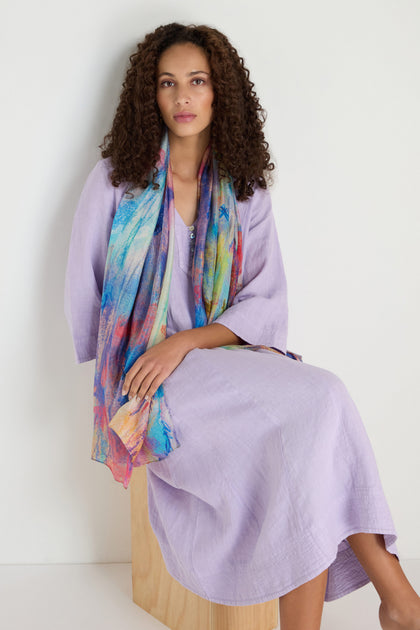 A woman with curly hair sits on a wooden block, wearing a light purple dress and the Soft Meadow Dreams Silk Scarf with a vibrant botanical print, posed against a plain white background.