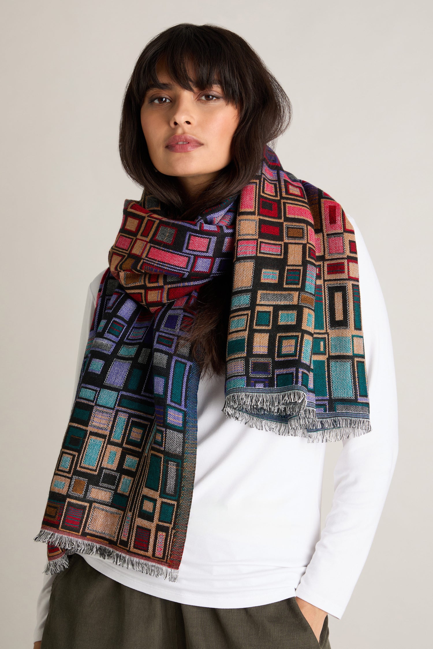 A person wears a white shirt and the Tesserae Scarf, which features a vibrant multicolored geometric pattern and fringe edges, adding a touch of contemporary flair.