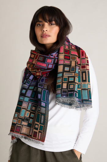 A person wears a white shirt and the Tesserae Scarf, which features a vibrant multicolored geometric pattern and fringe edges, adding a touch of contemporary flair.