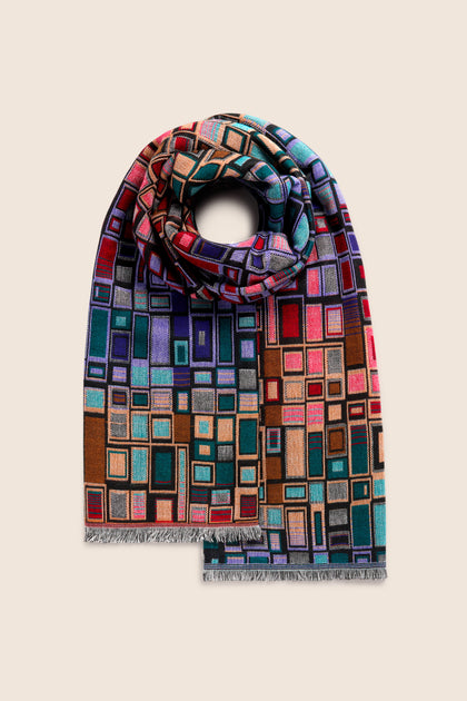 The Tesserae Scarf, rolled up and displayed on a plain beige background, features a vibrant abstract geometric pattern with squares and rectangles that add a touch of contemporary flair.