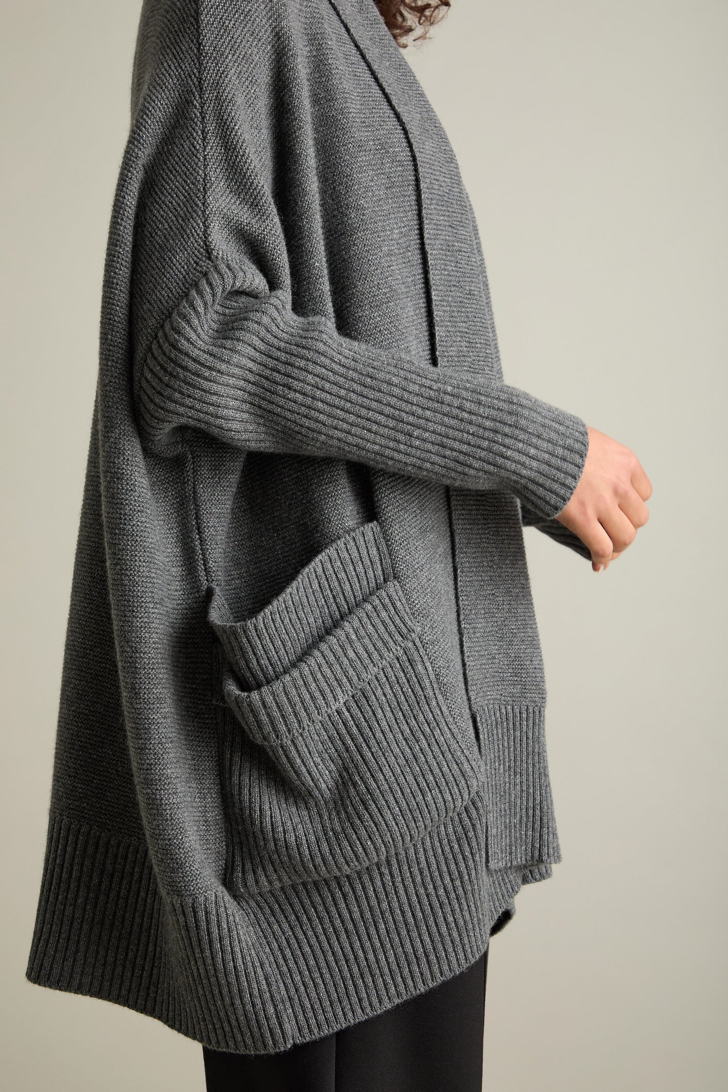 A person wearing the Pocket Detail Rib Cardi in gray, a wool-blend open-front cardigan featuring ribbed cuffs and pocket details, holds an unseen object against a plain background.