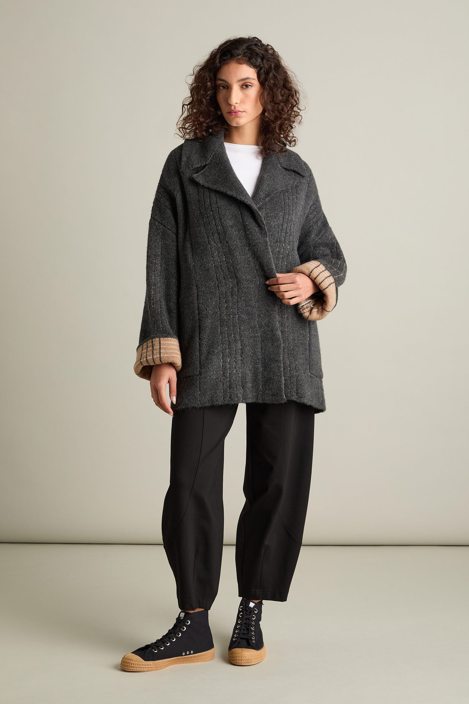 A person with curly hair wears a stylish Check Detail Jacket made from a wool blend in gray, paired with black pants and black sneakers, posing elegantly against a plain background.