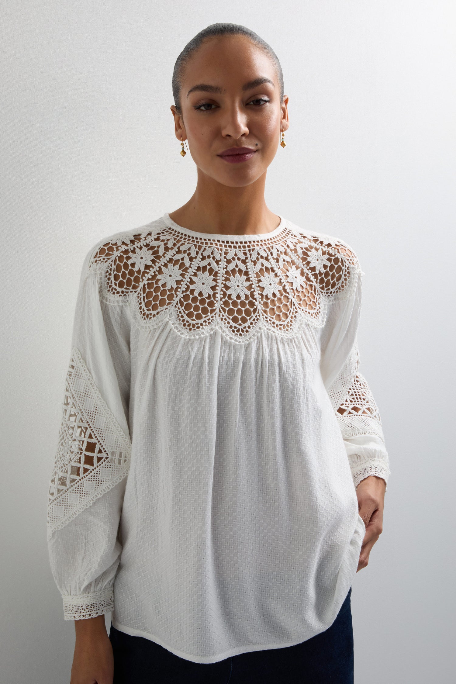 A person poses softly against a plain background, wearing the Floral Lace Trim Top, style KNT6038-FLA, featuring intricate patterns.