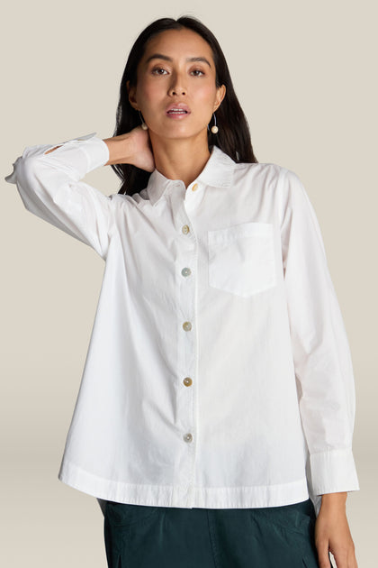 A person wearing a white Curved Hem Shirt made from Indian Cotton, featuring long sleeves and a pocket, is standing against a neutral background, with one hand touching their neck.