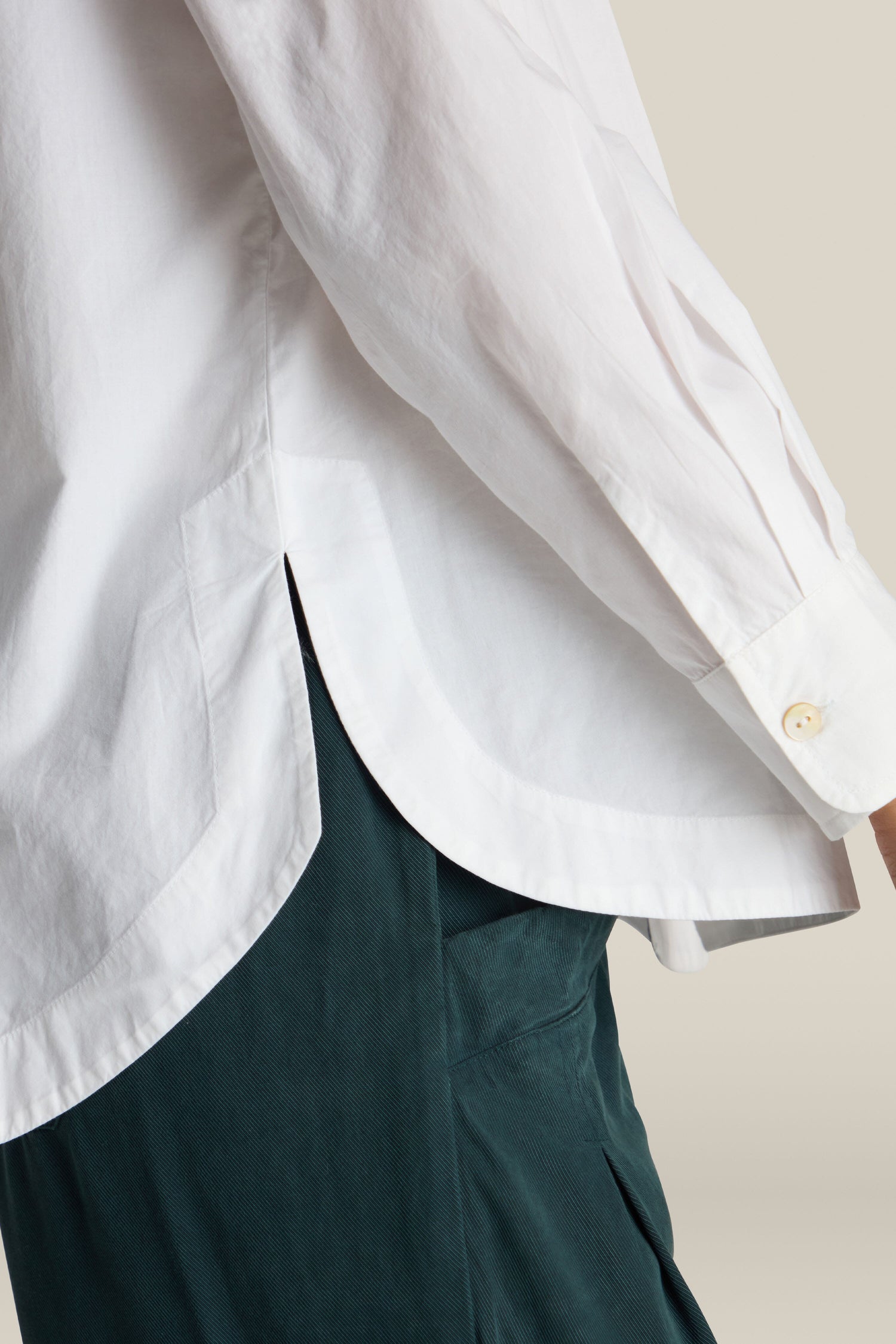 A close-up of a person wearing a Curved Hem Shirt with rolled-back cuffs and dark green pants. The photo captures the side view of the shirt's curved hem and a small part of the pants.