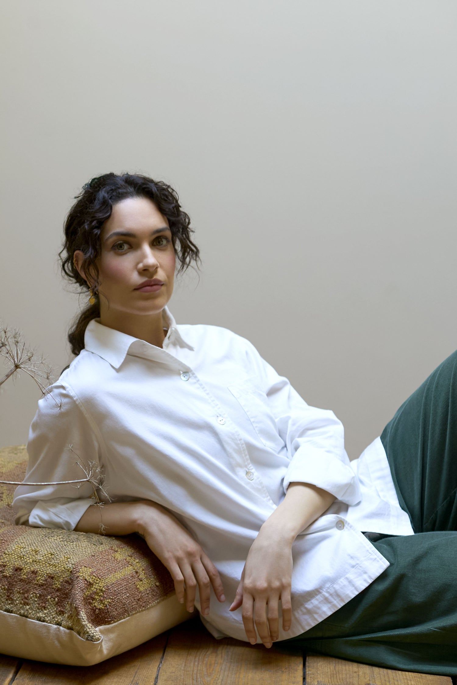 A woman with dark curly hair wearing a white Curved Hem Shirt with long sleeves and green pants lounges on a cushion against a beige background.