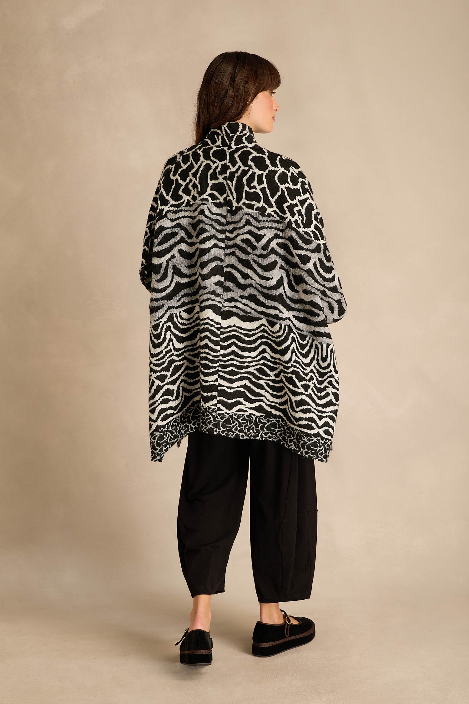 A person stands with their back to the camera, wearing the Abstract Jacquard Merino Wool Jacket with a bold black and white pattern, paired with black pants and black shoes, set against a neutral background.