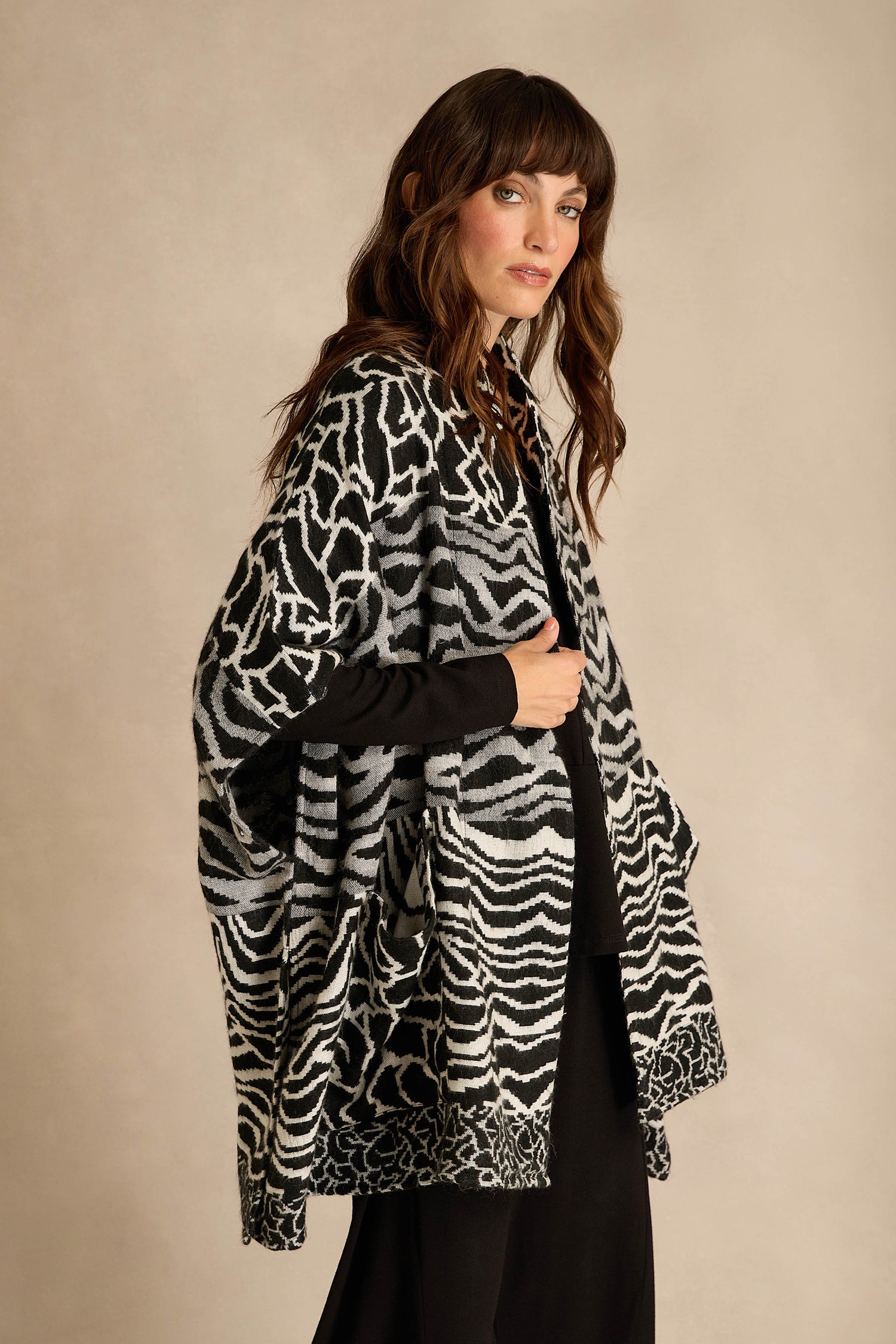 A woman wearing a black dress and the Abstract Jacquard Merino Wool Jacket, featuring a monochrome black-and-white animal print, stands against a beige background and looks at the camera.