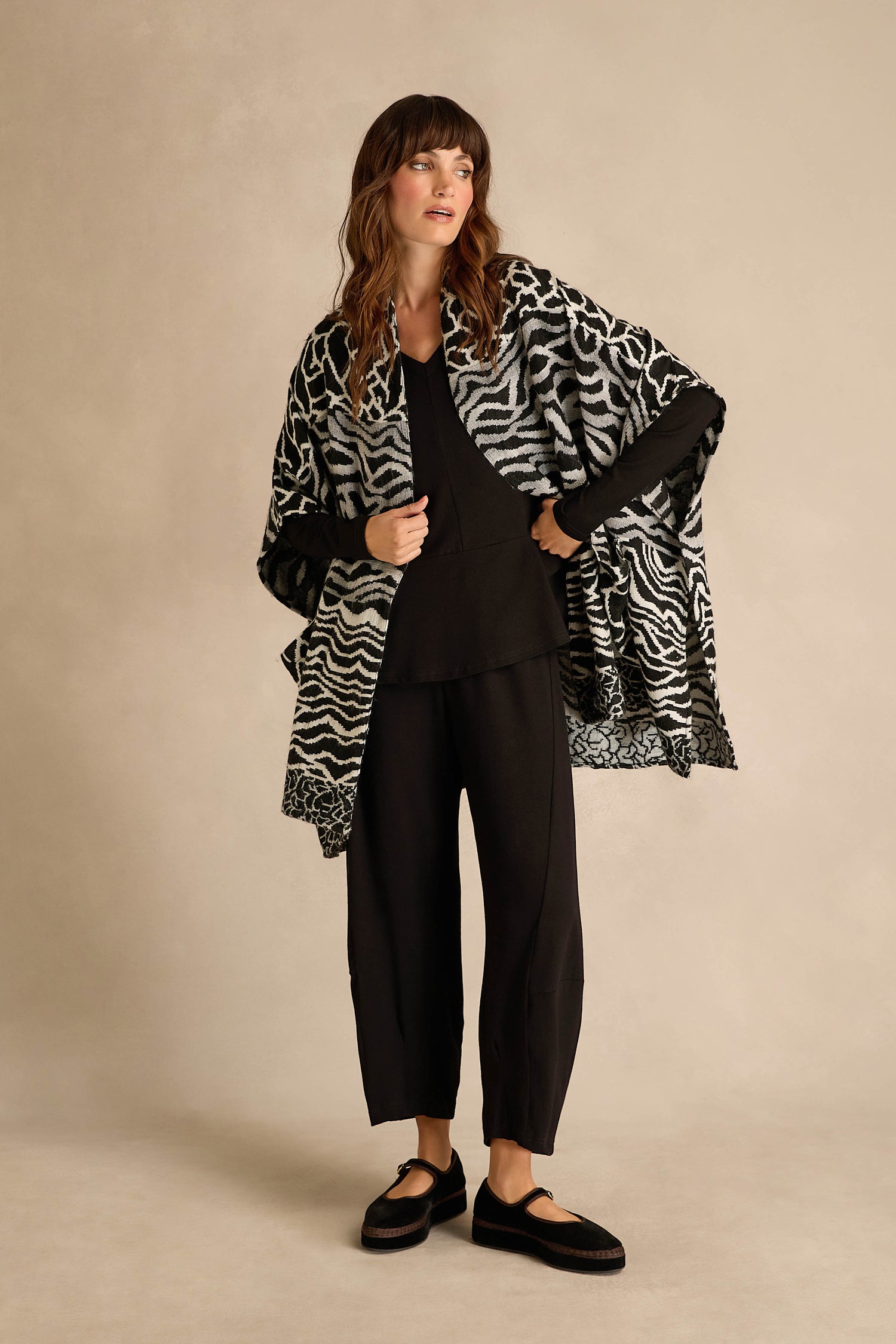 A woman stands in a studio wearing a black outfit topped with the Abstract Jacquard Merino Wool Jacket, which showcases a chic monochrome pattern. Her look is complemented by a black and white zebra-patterned shawl and black shoes.