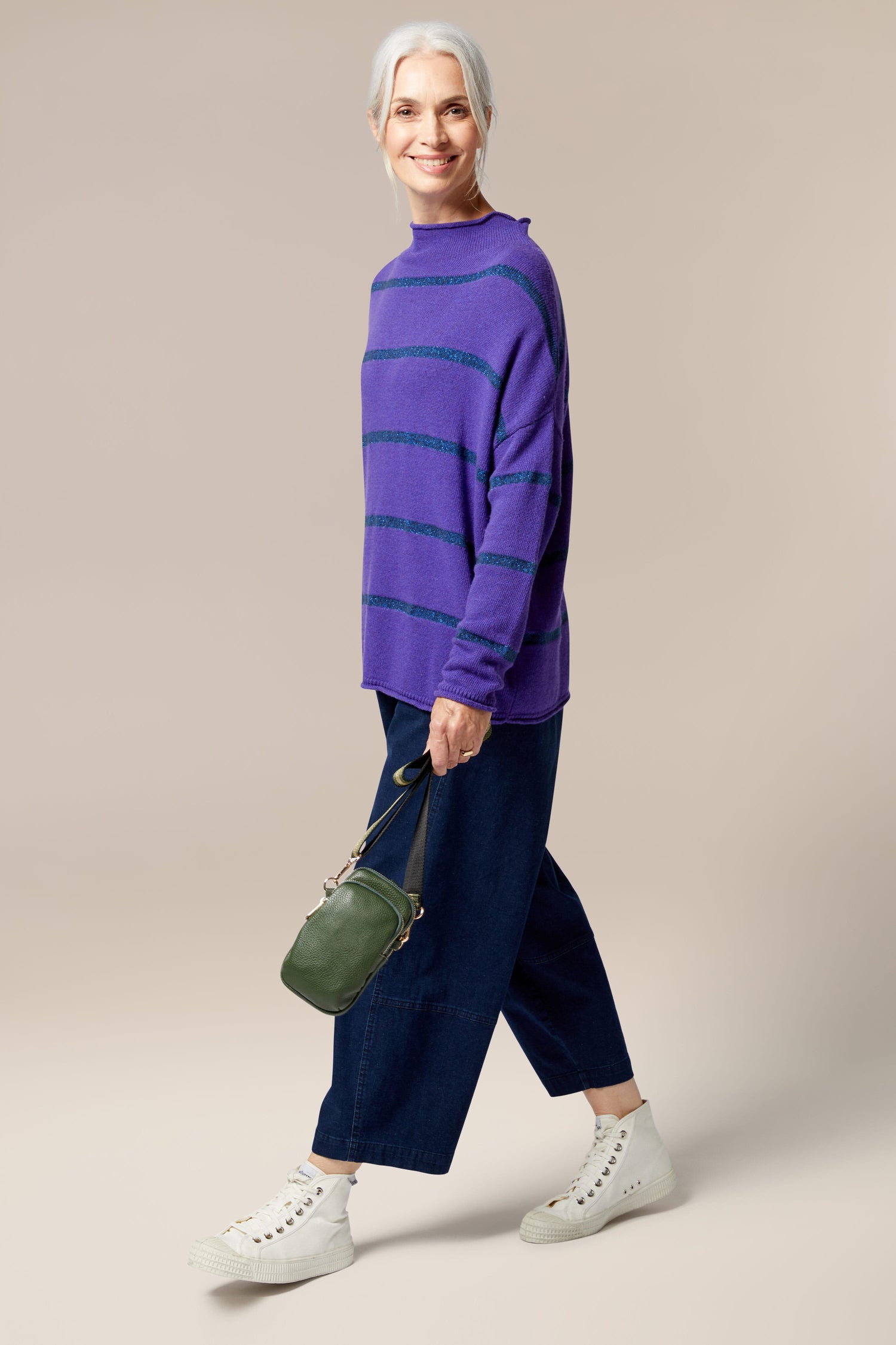 An older woman showcasing style-led designs in a purple sweater and wide leg pants by Kris-Ana, carrying a Mini Crossbody Bag.