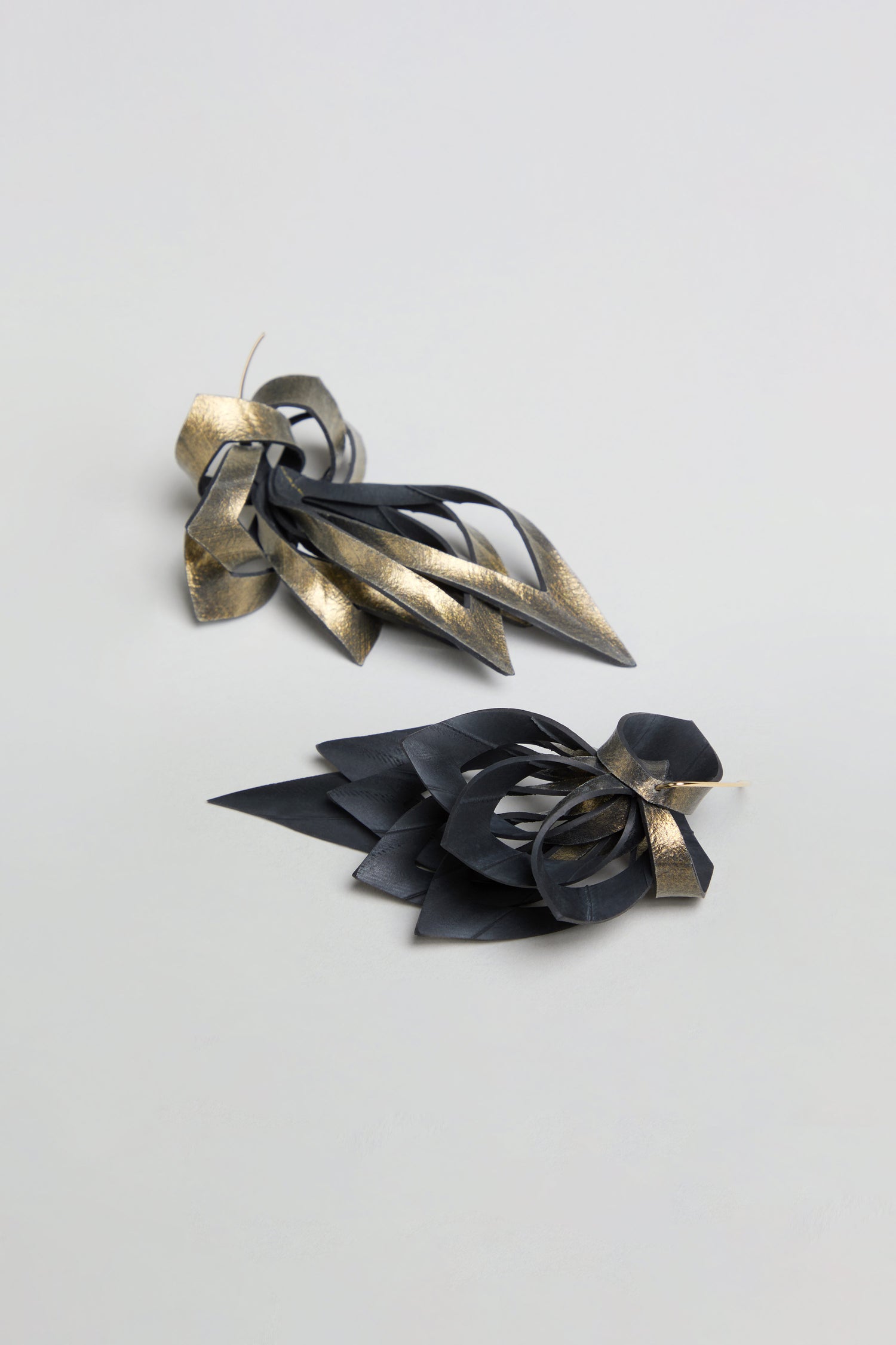 Two abstract, black and gold sculptural objects—the Gold Flipped Leaves Earrings—crafted from folded material reminiscent of reclaimed bicycle inner tube, each featuring pointed shapes and looped elements, are placed on a plain light grey surface.