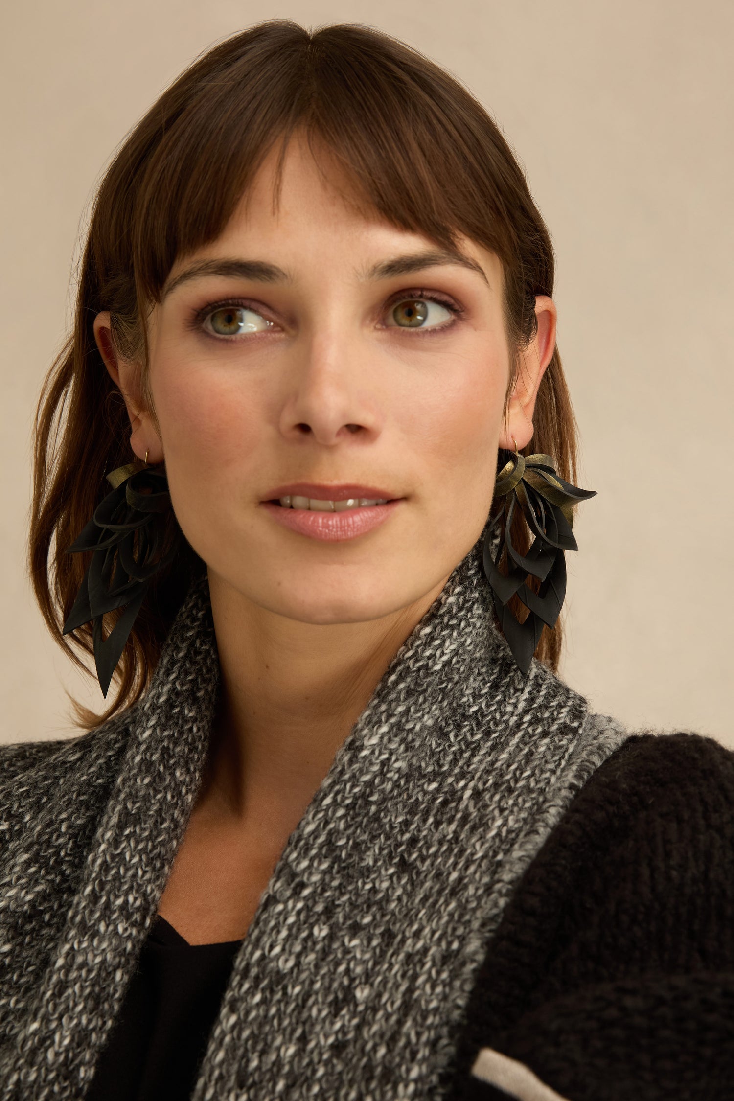 Woman with straight brown hair wearing Gold Flipped Leaves Earrings, a black top, and a textured gray sweater, looking slightly to her left against a neutral background.