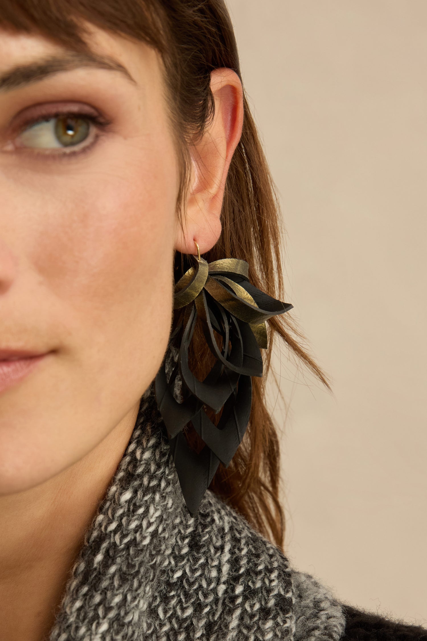 Close-up of a woman wearing the Gold Flipped Leaves Earrings—large, layered black and gold leaf drop earrings crafted from reclaimed bicycle inner tubes—paired with a gray knit scarf. Only the left side of her face is visible.