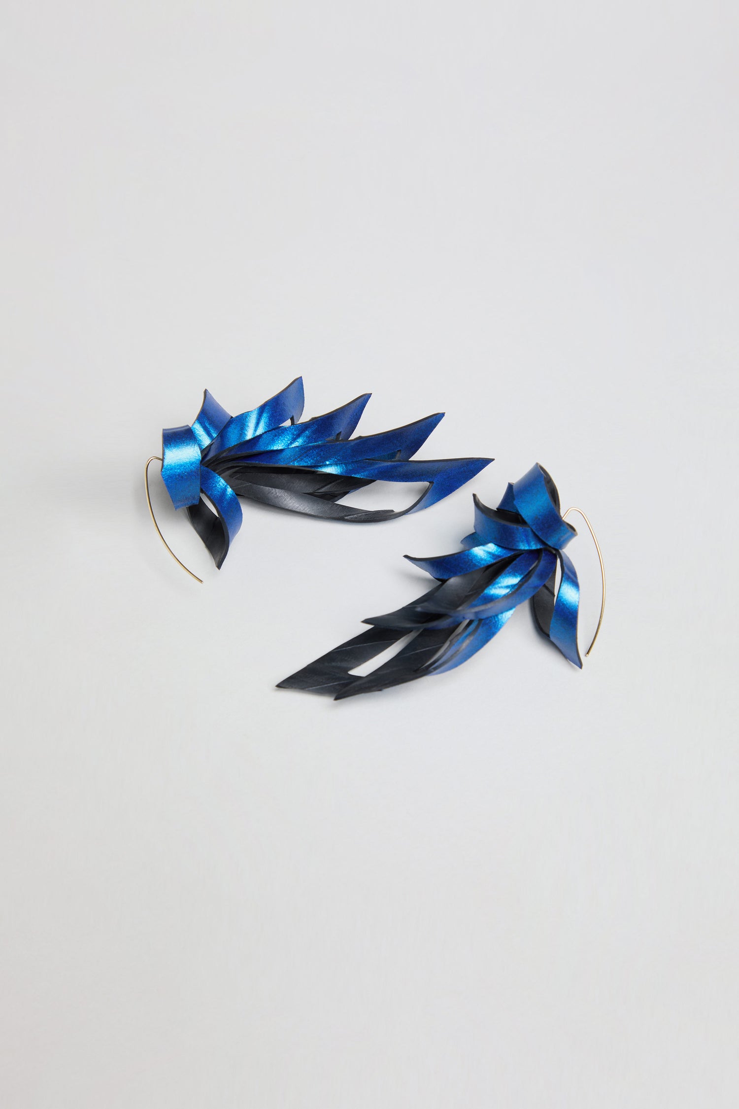 Introducing the Metallic Blue Flipped Leaves Earrings: a pair of feather-shaped earrings in blue and black with a metallic sheen, expertly crafted from reclaimed bicycle inner tubes. Displayed on a plain white background, these upcycled earrings add a distinctive, sustainable twist to classic feathered leaf designs—blending eco-friendly materials with bold style.