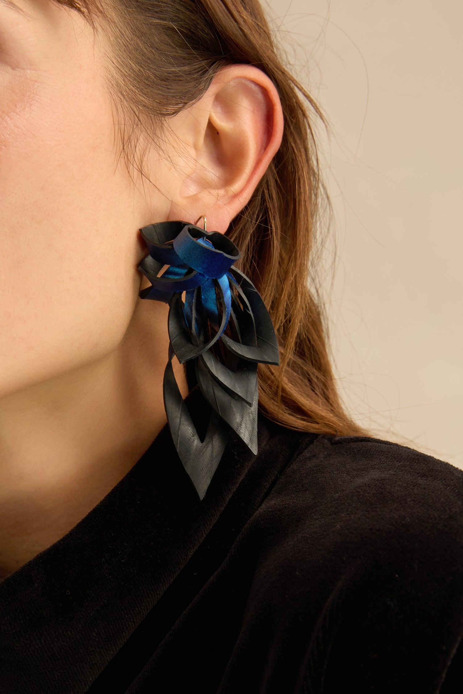 Close-up of a person wearing the Metallic Blue Flipped Leaves Earrings—striking, expertly crafted pieces made from reclaimed bicycle inner tube material and shaped like layered leaves in geometric black and blue. The wearer’s brown hair and black top are also visible.