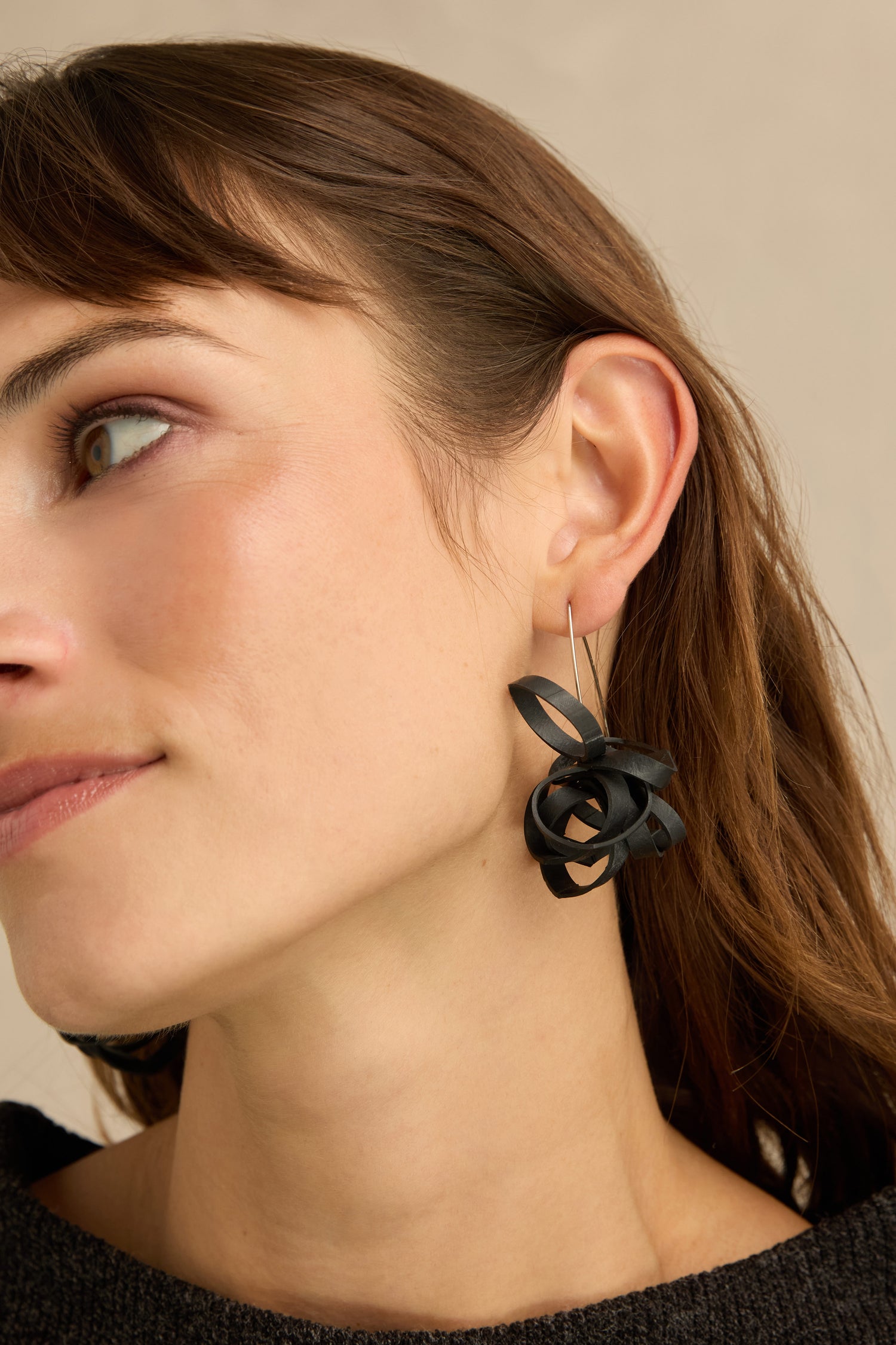 A woman with brown hair is wearing a Black Pom Pom Earring, a large abstract accessory crafted from upcycled bicycle inner tube jewelry. Shaped like a twisted ribbon, the earring is visible on her left ear as she looks slightly to the left, showing only the left side of her face.