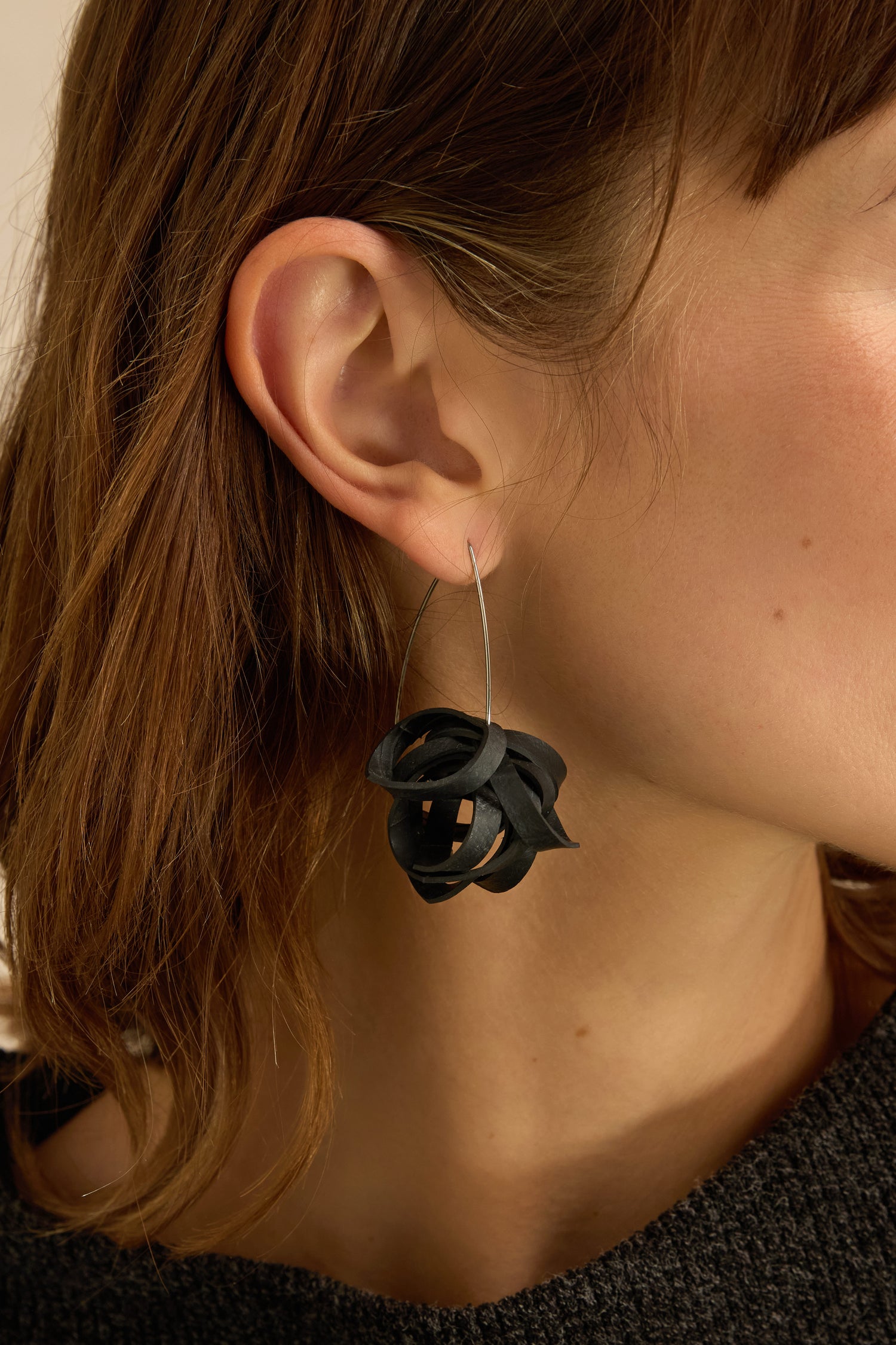 Close-up of a person with light skin and brown hair wearing Black Pom Pom Earrings—large, abstract-shaped accessories crafted from upcycled bicycle inner tubes, featuring a thin metal hoop.