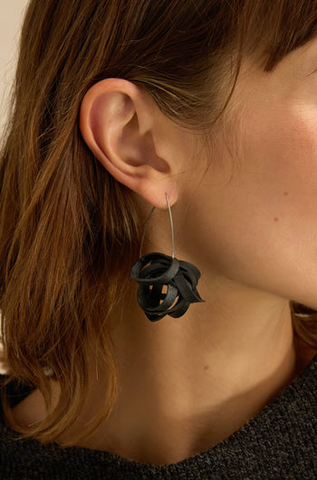 Close-up of a person with light skin and brown hair wearing Black Pom Pom Earrings—large, abstract-shaped accessories crafted from upcycled bicycle inner tubes, featuring a thin metal hoop.