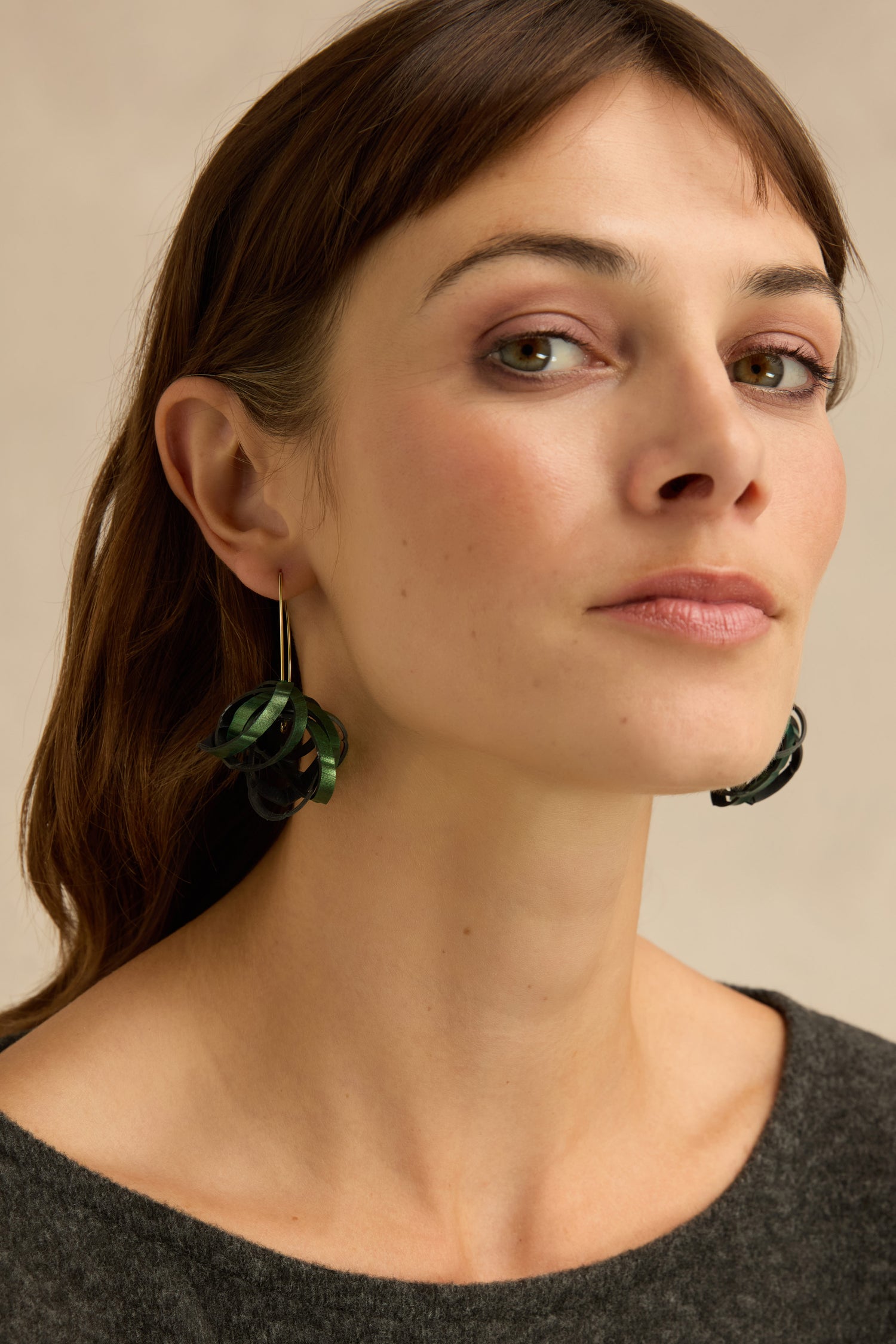 Woman with straight brown hair wearing Metallic Green Pom Pom Earrings made from reclaimed bicycle inner tube in a dark green, drop style, paired with a grey top, looking slightly upward with a neutral expression against a beige background.