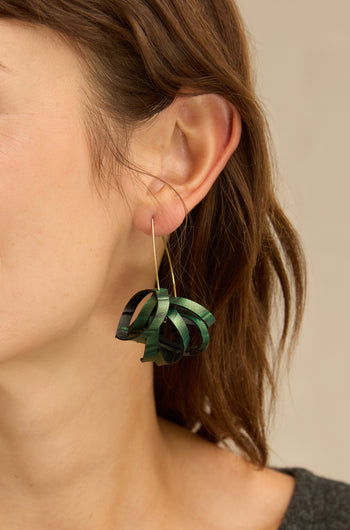 Close-up of a person wearing the Metallic Green Pom Pom Earrings, featuring a large, dark green, ribbon-like drop design with a gold hook. The earring hangs below the ear, and the person has shoulder-length brown hair.