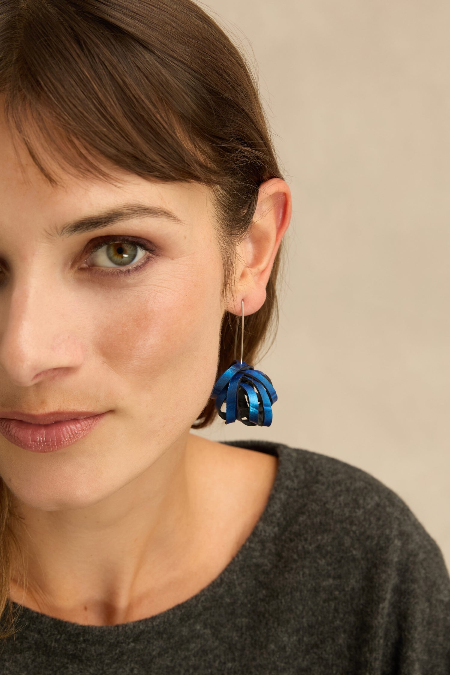A woman with light skin and brown hair, wearing a gray top and a pair of striking Metallic Blue Pom Pom Earrings, crafted from upcycled bicycle inner tubes, photographed against a neutral background.