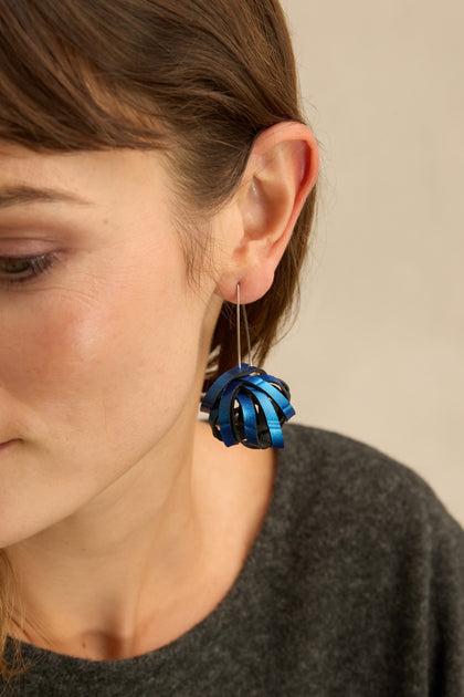 Close-up of a woman’s ear wearing Metallic Blue Pom Pom Earrings made from upcycled materials. She has straight brown hair and is dressed in a dark grey top, highlighting her unique taste for upcycled earrings.