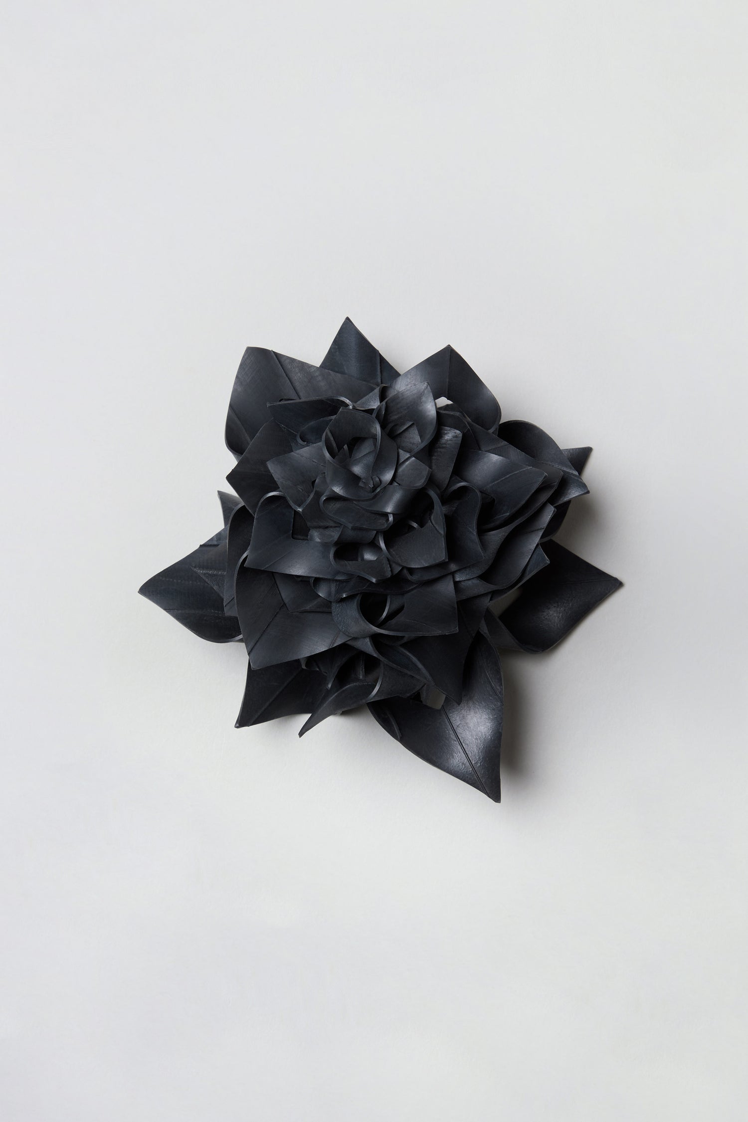 Presenting the Rose Corsage—a unique black flower with layered petals, expertly crafted from upcycled bicycle inner tubes to create a striking, rubber-like appearance. Displayed against a plain light background, this accessory highlights the creative charm and eco-friendly appeal of the Rose Corsage.