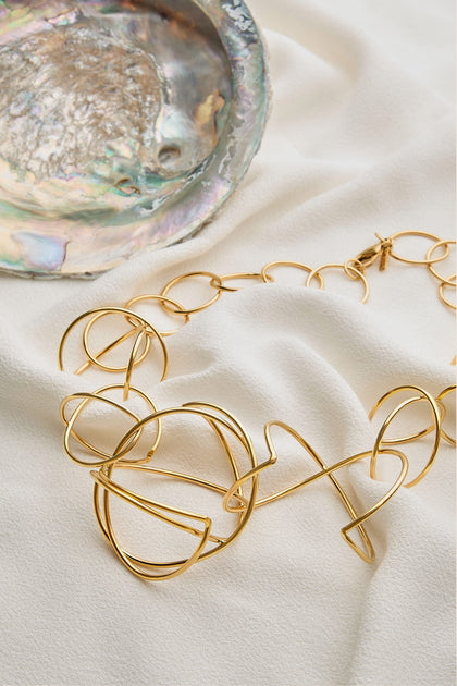 The Handmade Links Necklace, crafted from 24k gold plated brass and featuring contemporary abstract loops, is elegantly displayed on a white fabric beside an iridescent decorative shell, highlighting the sophistication of handmade jewelry.