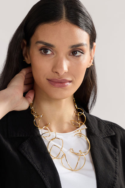 A person with long dark hair wearing a black blazer over a white shirt and the Handmade Links Necklace, featuring an abstract gold design.