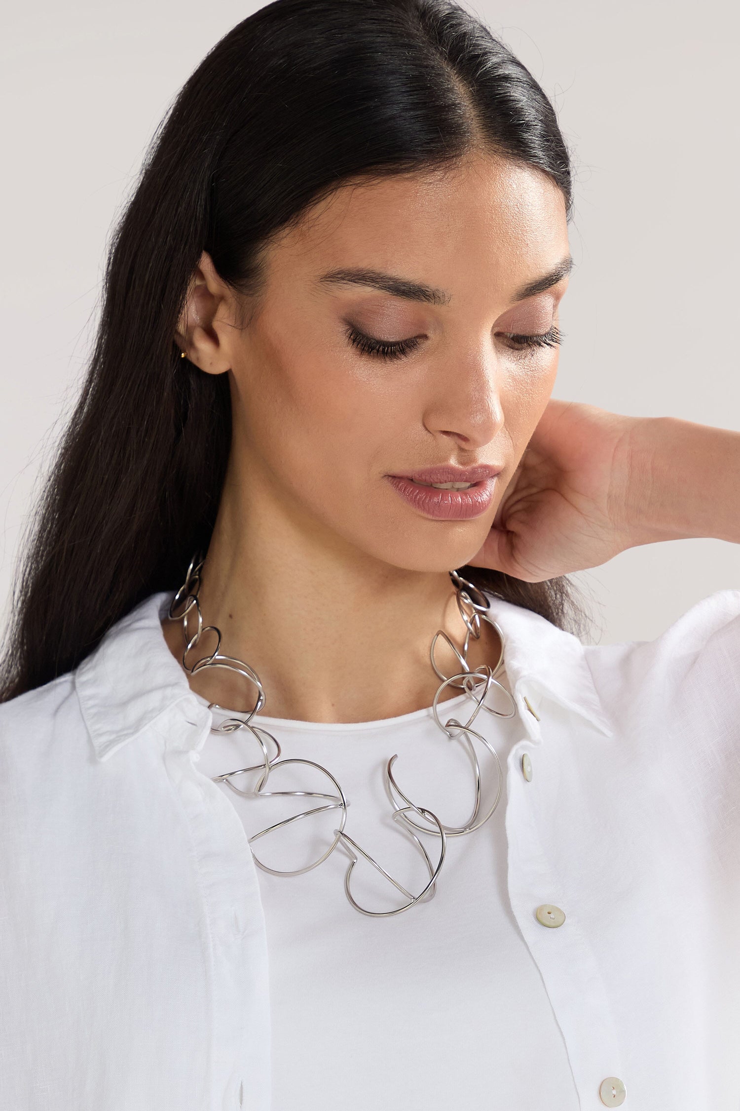 A woman with long dark hair wearing a white button-up shirt and a large, intricate Handmade Links Necklace looks down with one hand behind her head.