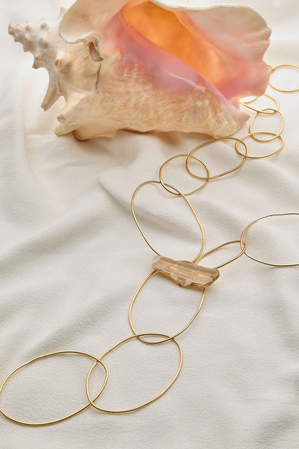 The Handmade Crystal Quartz Rings Necklace, characterized by substantial oval links and a central quartz pendant, is elegantly displayed on a white fabric beside a large conch shell.