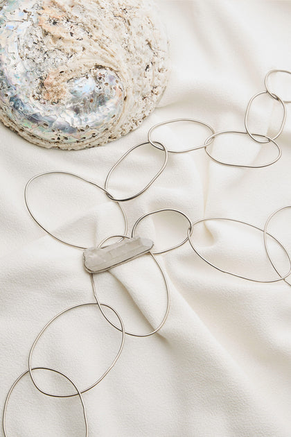 The Handmade Crystal Quartz Rings Necklace, featuring large, interlocking loops and a central white quartz pendant, is displayed on a white fabric background next to a textured shell.