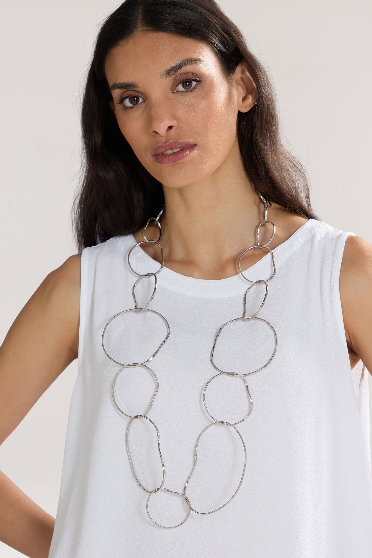 A woman with long dark hair wears a white sleeveless top and the Handmade Silver Hoops Necklace by a renowned Greek designer, featuring large oval links. She is standing against a plain background.