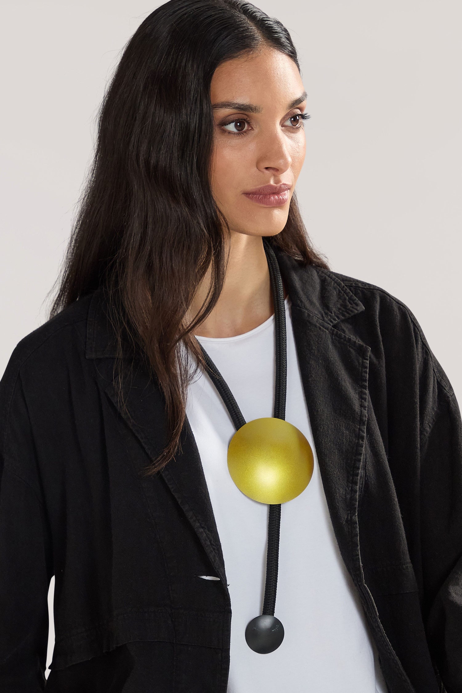 A woman with long dark hair, wearing a black jacket and white shirt, showcases the Handmade Aphelion Necklace – a large, round yellow pendant designed by a Greek jewellery designer, adding a bold contemporary touch with its handmade craftsmanship.