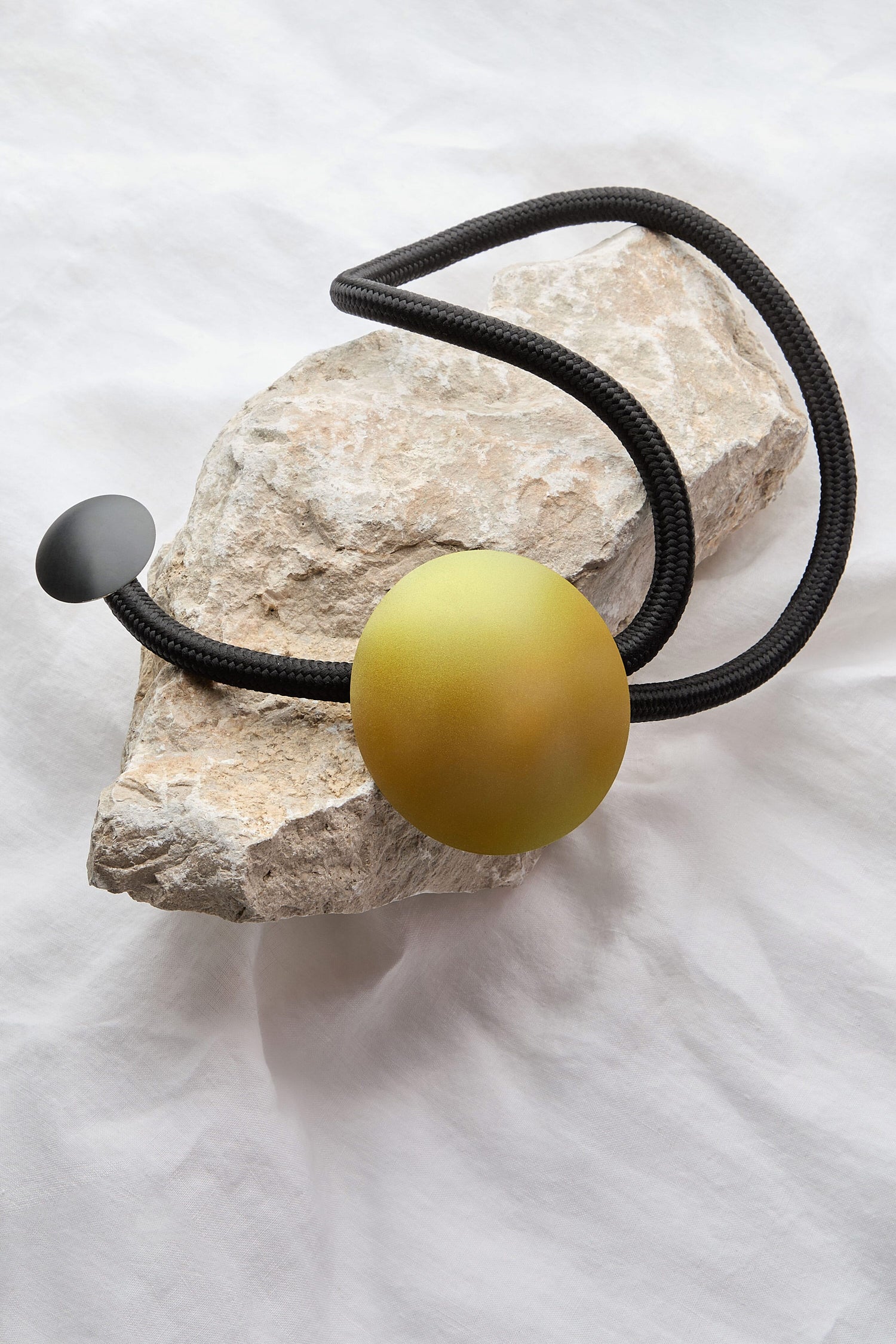 The Handmade Aphelion Necklace, featuring a bold contemporary design with a black cord and a large green spherical pendant complemented by a smaller black disc, is elegantly arranged on a rough-textured rock.