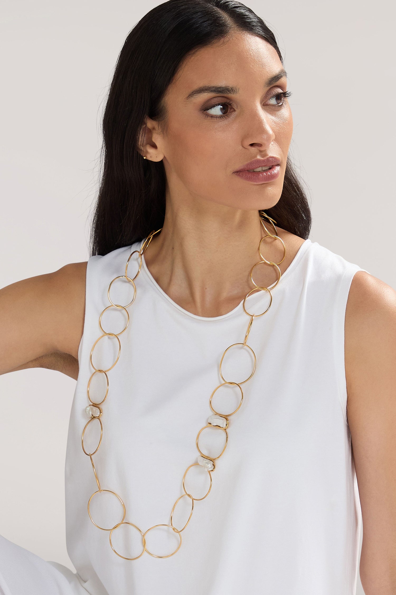 A woman with long dark hair wearing a white sleeveless top and the Handmade Pearl Hoop Necklace from a Greek jewellery designer sits against a light background, looking to her left.