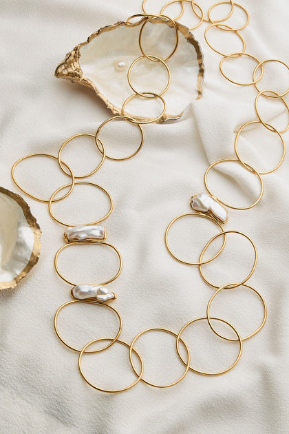 The Handmade Pearl Hoop Necklace is artistically arranged on a white fabric adorned with pearl accents. Two opened oyster shells, each cradling a pearl, lie beside it, showcasing the exquisite craftsmanship of a renowned Greek jewellery designer celebrated for their fine handmade creations.