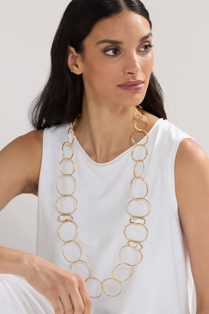 A woman with long dark hair is wearing a white sleeveless top and a Handmade Pearl Hoop Necklace, complemented by 24k gold plated brass hoops from a renowned Greek jewellery designer.