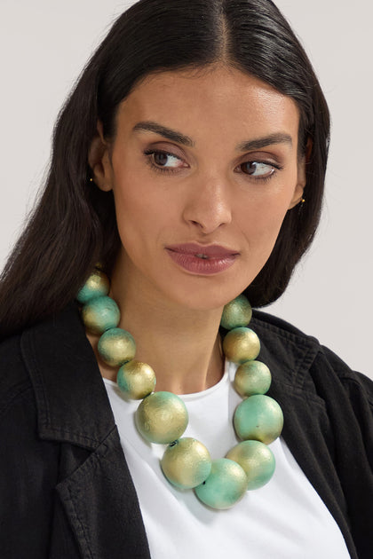 A woman with long dark hair wears a black jacket and the Handmade Short Painted Spheres Necklace, a bold piece featuring large green and gold beads. Crafted by a Greek designer, this necklace adds a touch of elegance as she looks to the side.