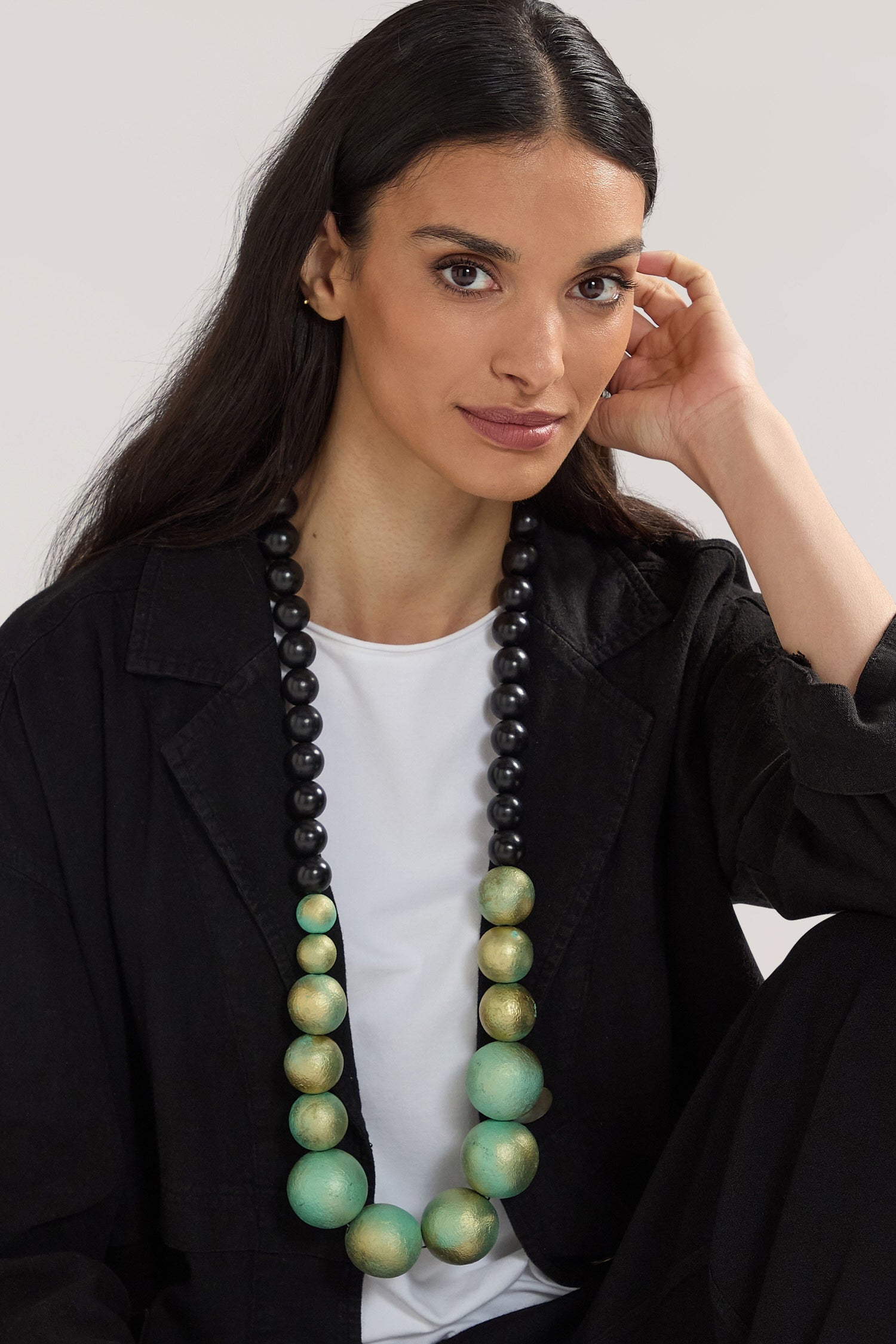 A woman with long dark hair, dressed in a black outfit, sits with her hand on her face while wearing the Handmade Painted Spheres Necklace, designed by a Greek jewellery designer, which features bold black and green beads.