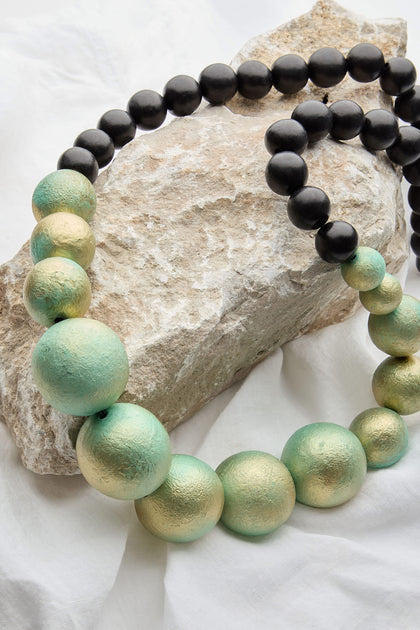 The Handmade Painted Spheres Necklace, crafted by a renowned Greek jewellery designer, features bold long necklaces adorned with black and green-gold beads. Displayed elegantly on a rock against a white fabric background, these handmade pieces capture timeless charm and sophistication.