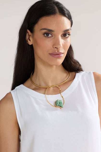 A woman with long dark hair is wearing a white sleeveless top and the Handmade Agate Hoop Necklace, featuring a striking 24k gold-plated brass choker adorned with a green stone pendant. She is standing against a plain background.