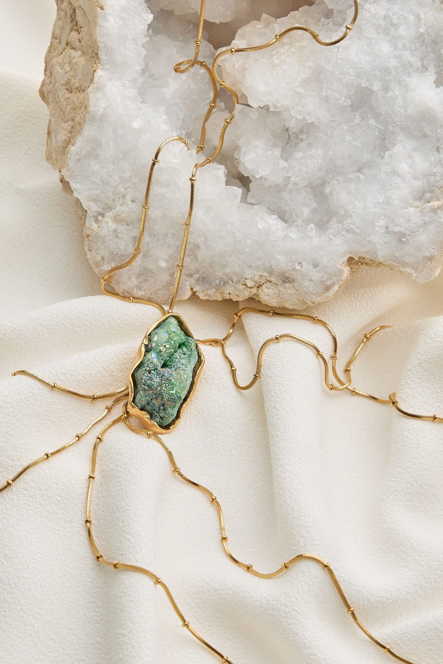 The Handmade Gold Plated Agate Pendant, featuring a green semi-precious Agate gemstone on a 24k gold plated brass chain, is elegantly displayed against a white textured rock and fabric backdrop, highlighting its exquisite craftsmanship.